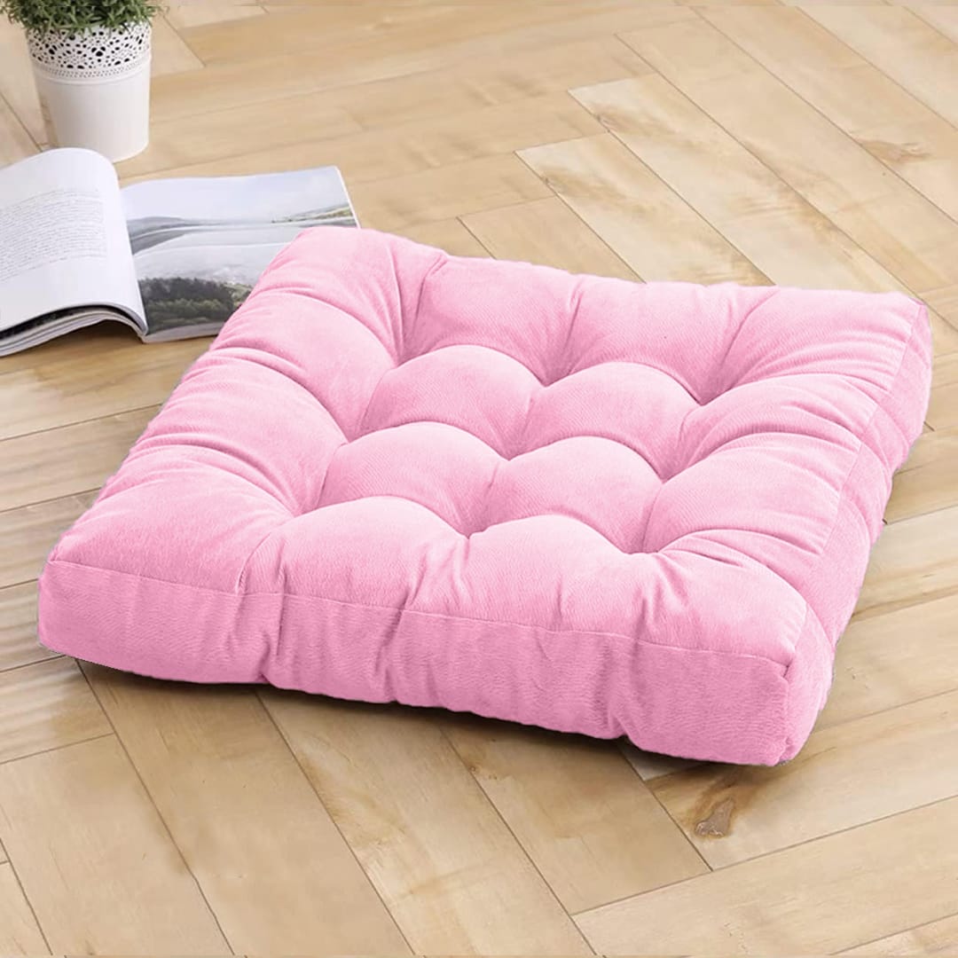 A pink square-shaped velvet floor cushion placed on a wooden floor, with a plant and a book beside it.